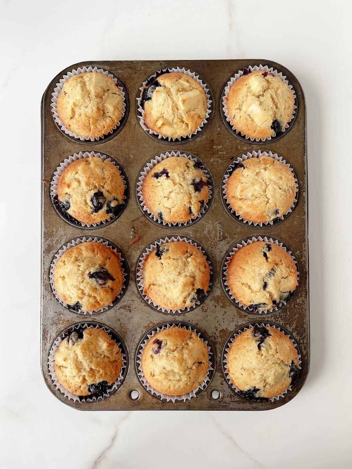 Top view of baked blueberry apple muffins in the metal muffin tin on a white marble surface.