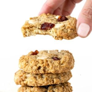 Close up of hand lifting a bitten oatmeal cookie with white chips and cranberries from a stack. White background.