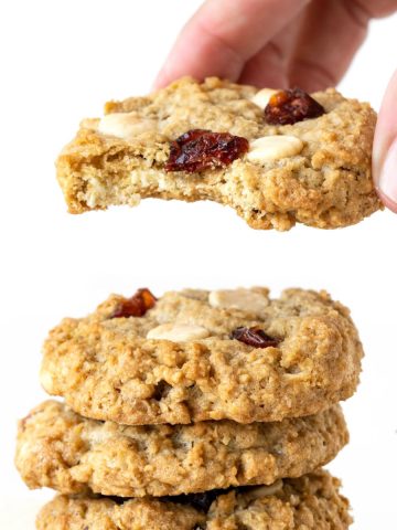 Close up of hand lifting a bitten oatmeal cookie with white chips and cranberries from a stack. White background.