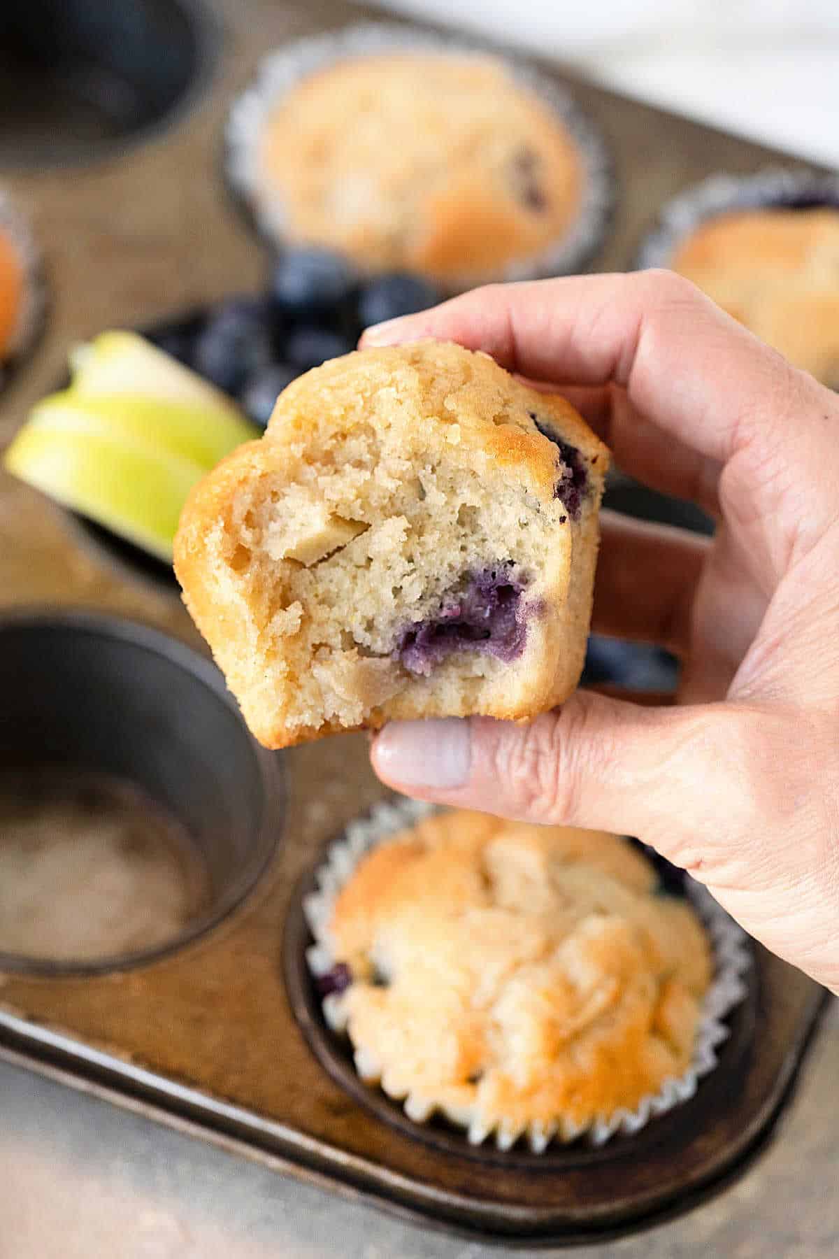 Eaten blueberry apple muffin being held over metal muffin tin with more muffins and fruit pieces.
