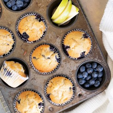 Metal muffin pan with apple blueberry muffins and fruit pieces. White cloth, beige surface.