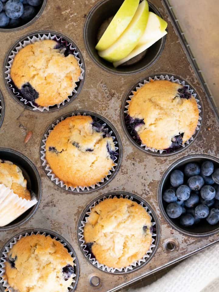 Metal muffin pan with apple blueberry muffins and fruit pieces. White cloth, beige surface.