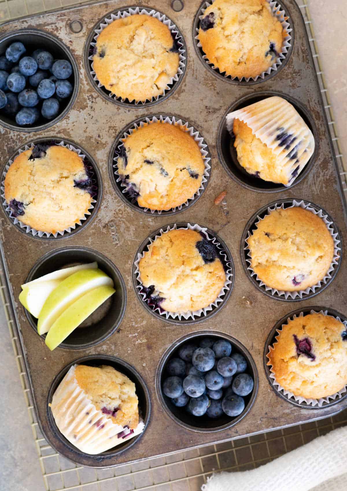Top view of metal muffin pan with apple blueberry muffins and pieces of said fruit.