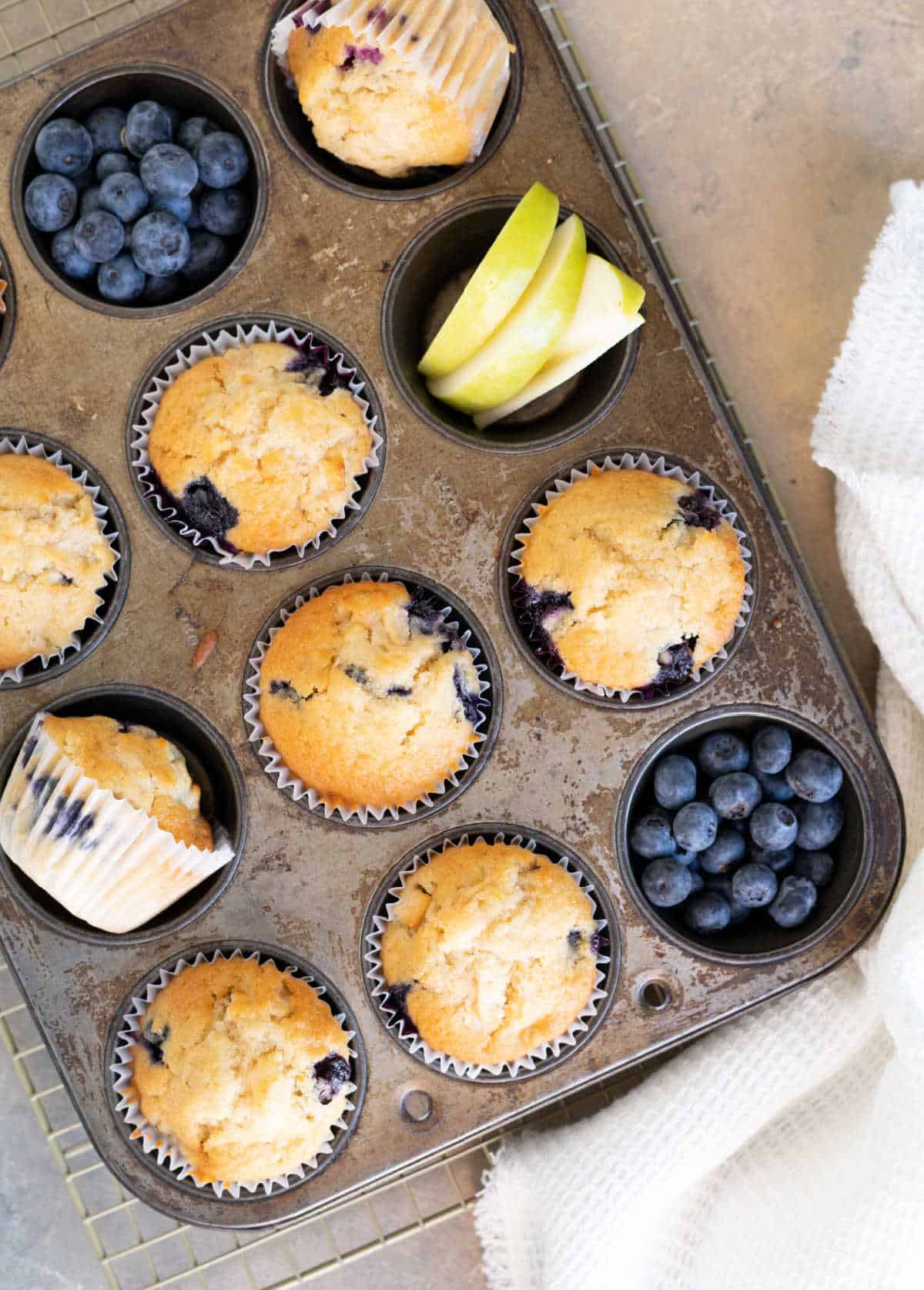 Apple blueberry muffins in the metal muffin tin with fruit around it. A white cloth and beige surface. Top view. 