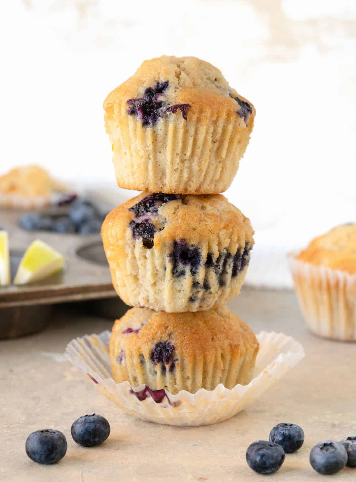 Stack of three blueberry apple muffins on a beige surface with white background. A metal muffin tin and loose blueberries.