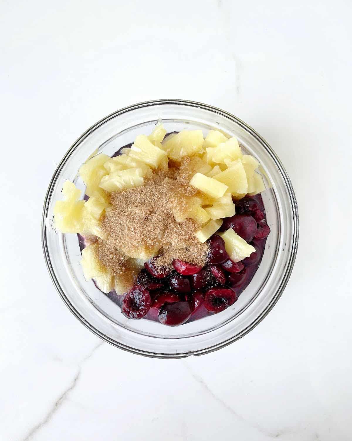 Cherries, pineapple chunks, brown sugar, and lemon juice in a glass bowl on a white marble surface.