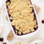 Partial view of white baking dish with cherry and pineapple cobbler on a wooden board over a white surface.
