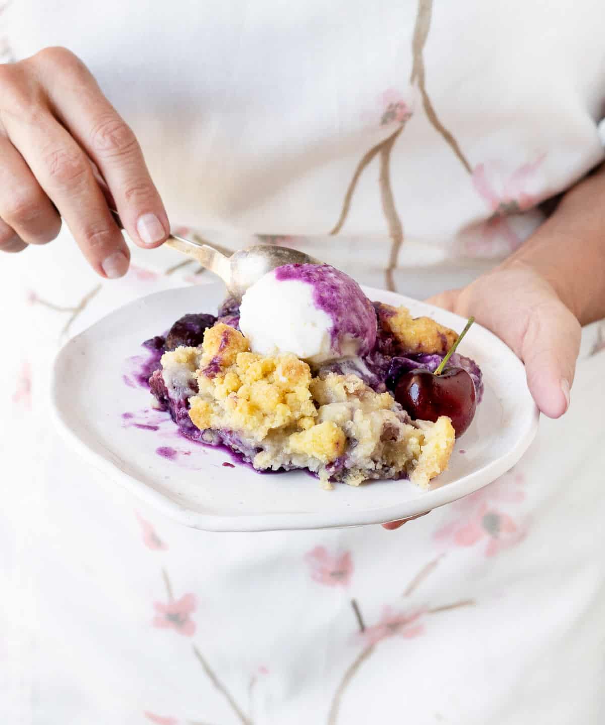 Spooning a white plate with cherry pineapple dump cake serving and ice cream. White and cherry blossoms apron in the background. 