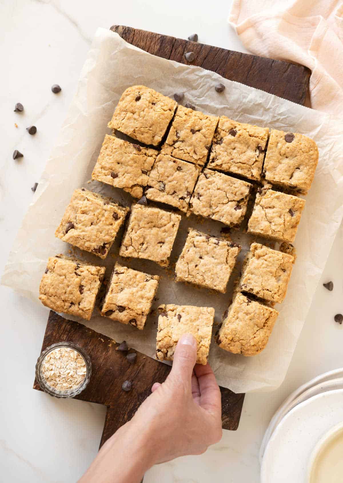 Overview of chocolate chip oatmeal bars on a beige parchment paper and dark wooden board. Light colored surface. Hand grabbing one bar.