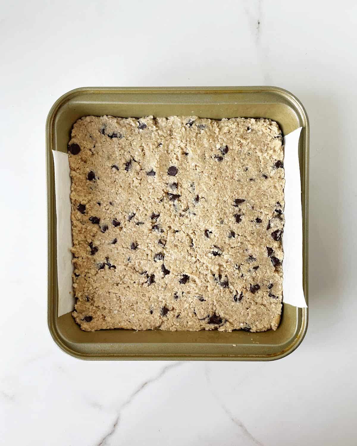 Raw chocolate chip cookie dough patted in a square metal pan on a white marble surface.