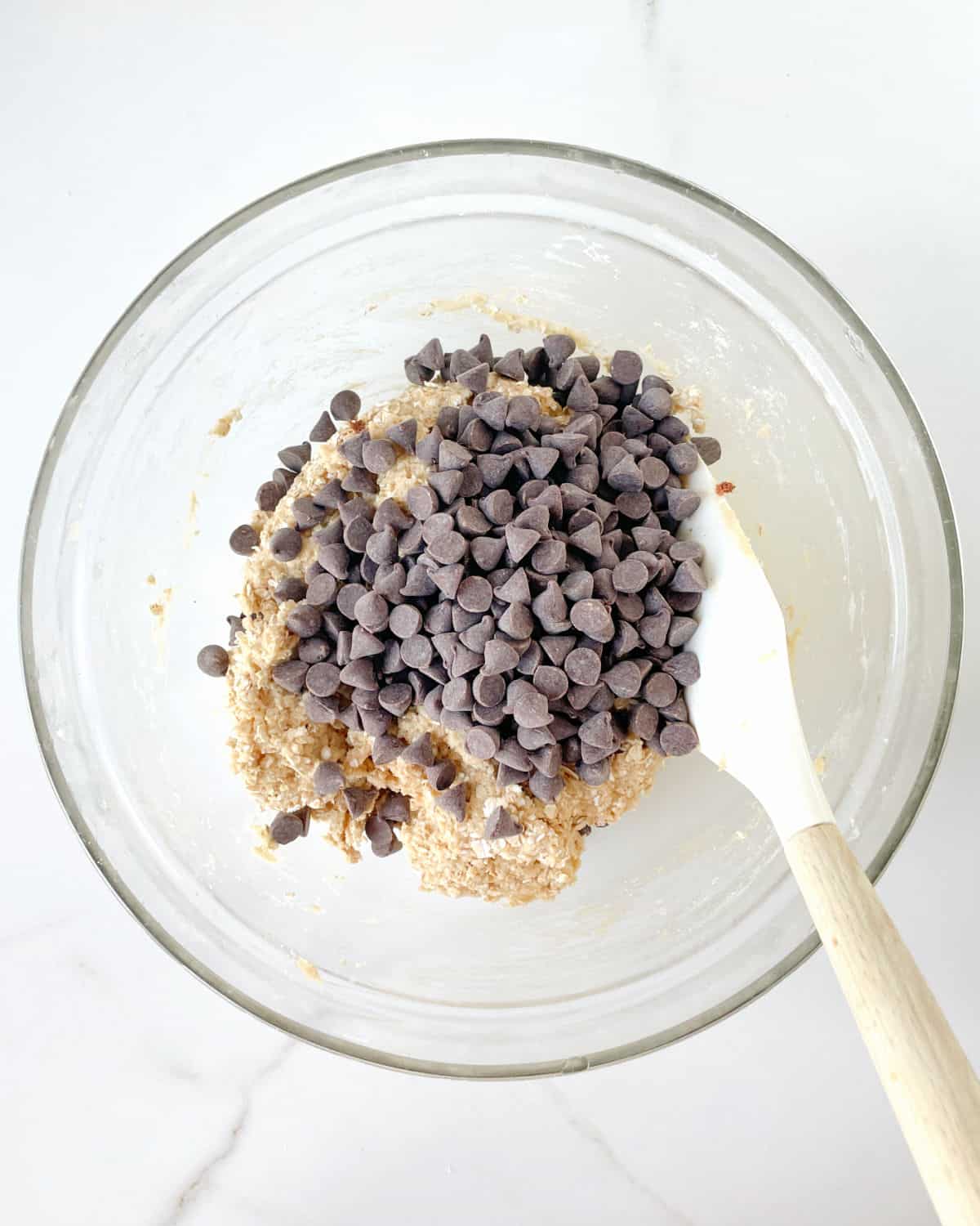 Chocolate chips added to cookie dough in a glass bowl with a white silicon spatula. White marble surface.