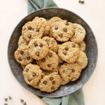 Close up of metal plate with piled chocolate chip oatmeal cookies. Green cloth beneath it. Light peach surface.