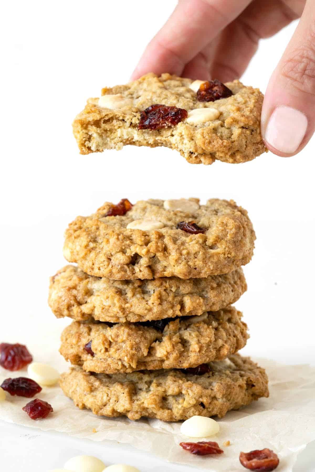Hand lifting bitten cranberry white chip cookie from a stack on a white surface and background.