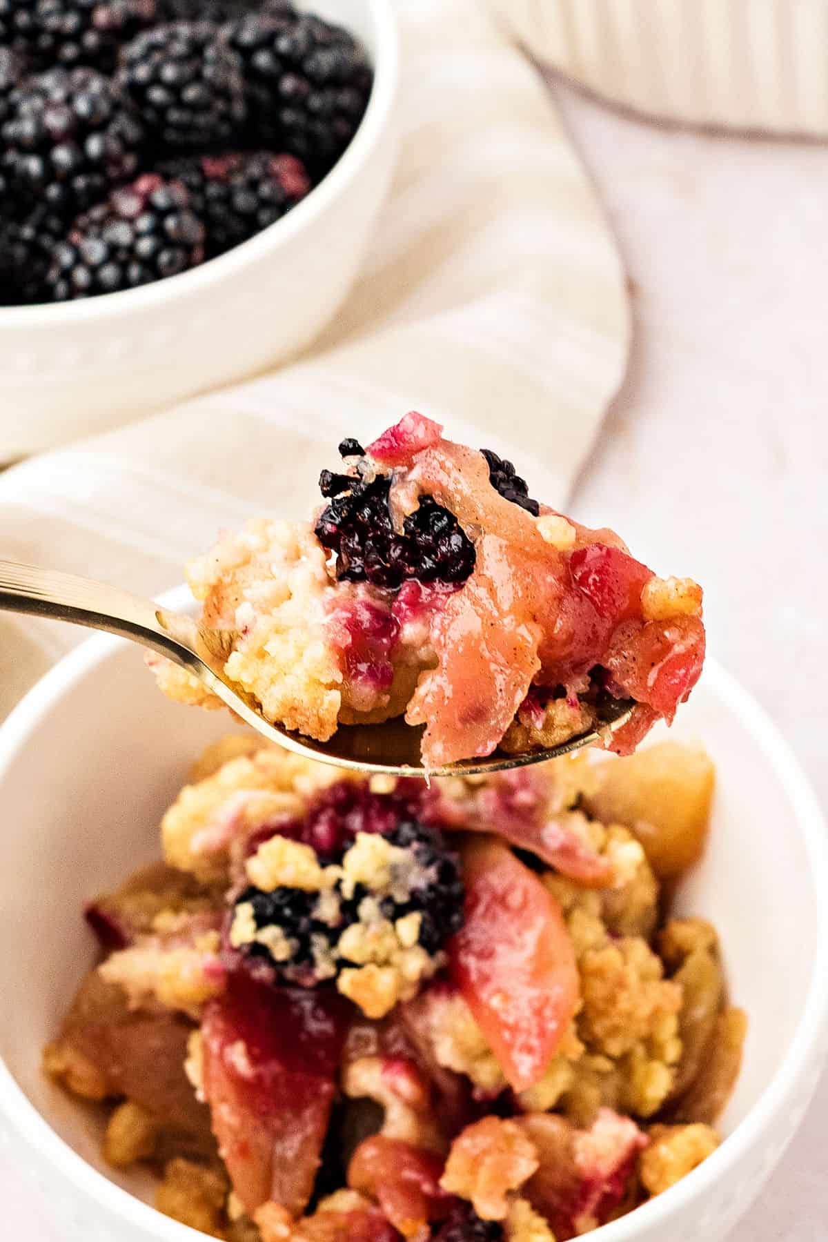 Spoon of apple blackberry crumble over served bowl. Bowl of fresh berries, whitish background.