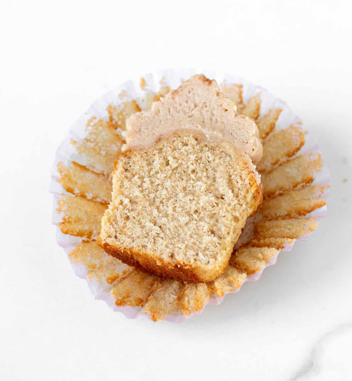 Halved cinnamon cupcake with frosting on an opened paper liner on a white marble surface. 