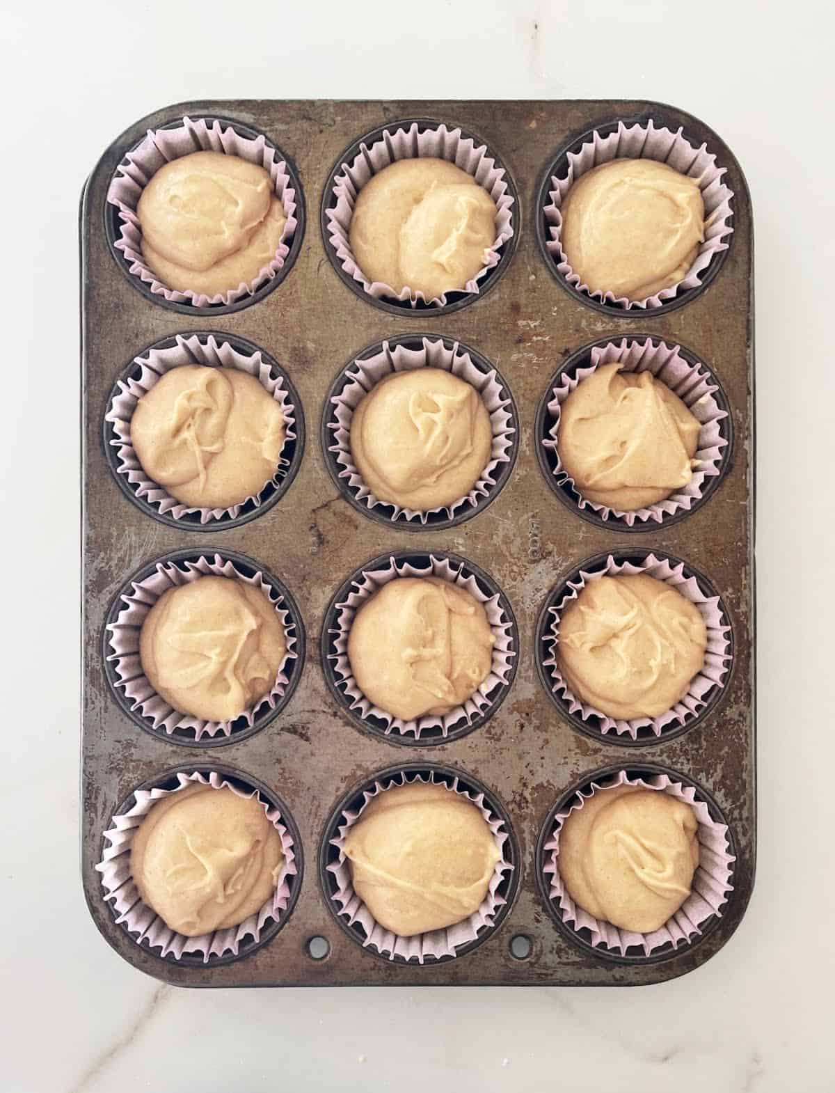 Muffin pan with cinnamon cupcake batter in paper liners. White marble surface.