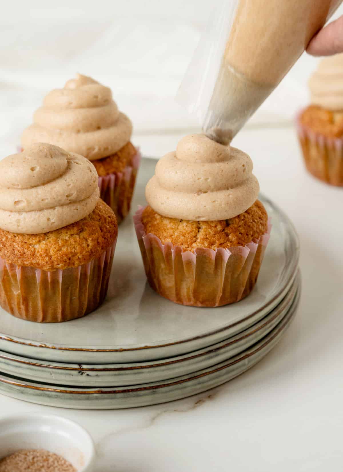 Using a piping bag to frost cinnamon cupcakes in paper liners on a stack of grey plates. White surface and background.