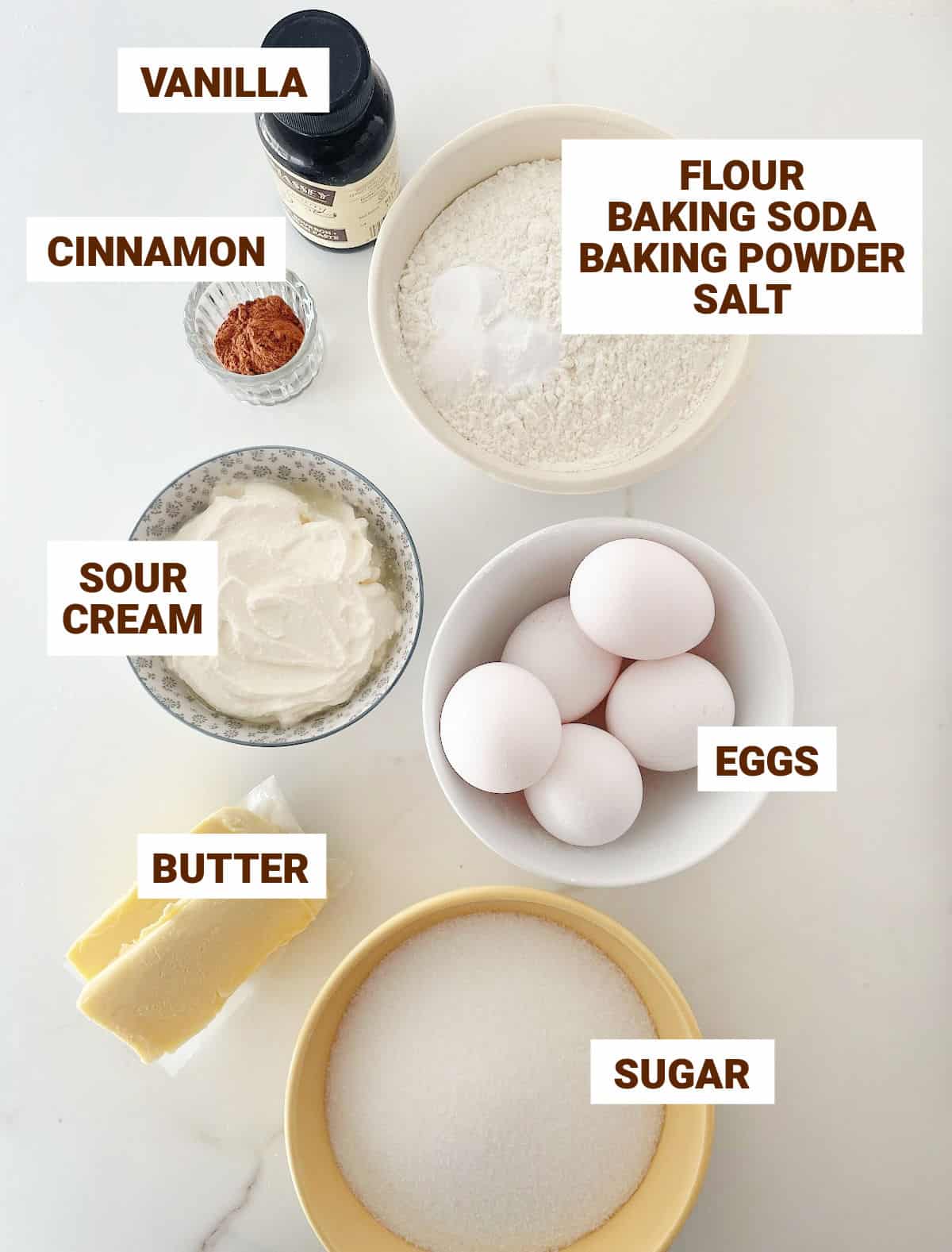 White marble surface with bowls containing ingredients for cinnamon sour cream cake including vanilla, flour mixture, sugar, eggs, butter, and vanilla.