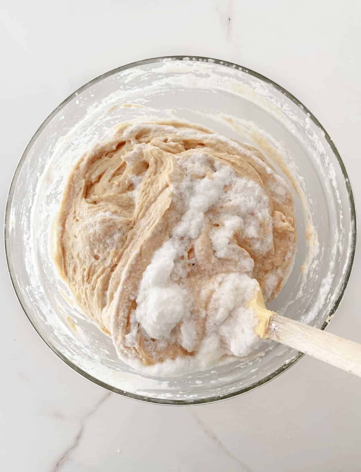 Glass bowl with cinnamon cake batter and whipped egg whites being mixed. White marble surface.