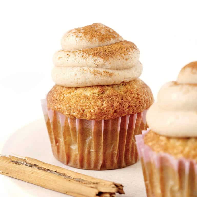Two frosted cupcakes and a cinnamon stick on a pinkish cake stand with white background.