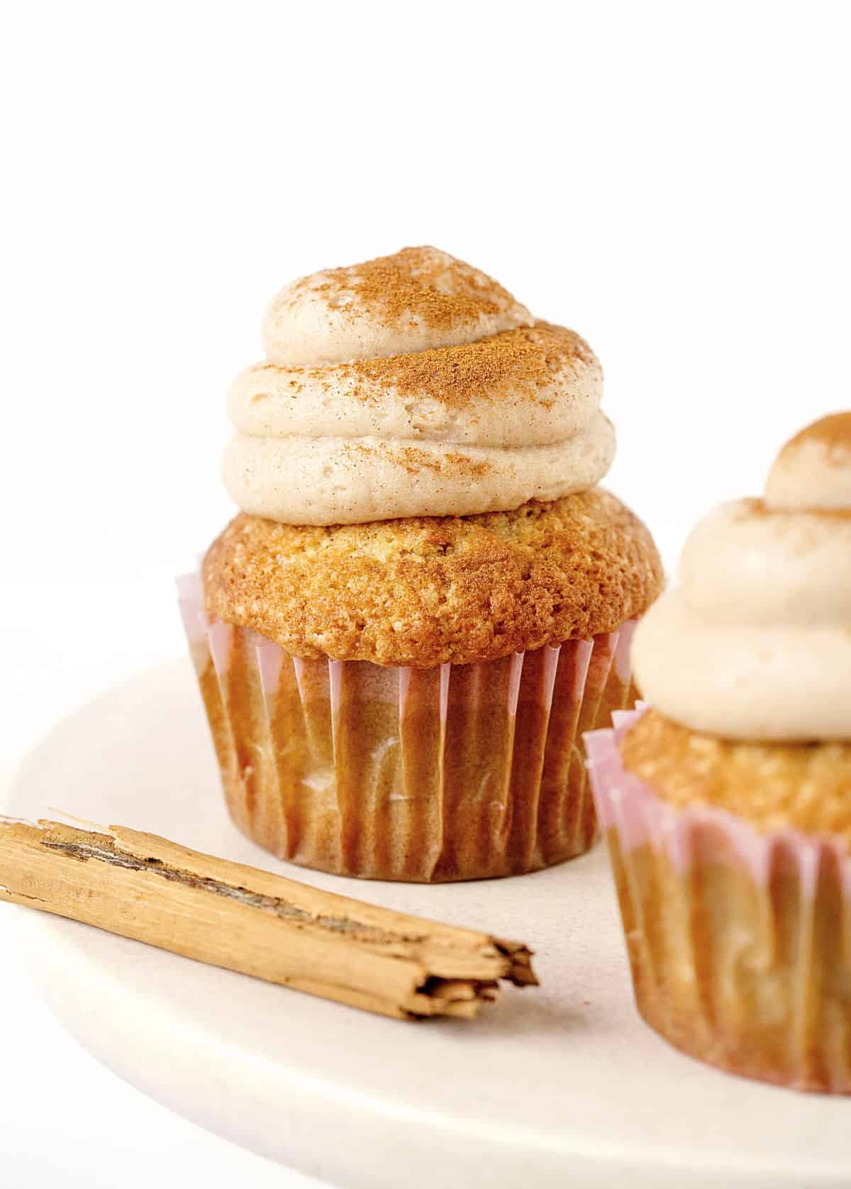 Beige pink cake stand with two frosted cupcakes and a cinnamon stick. White background.