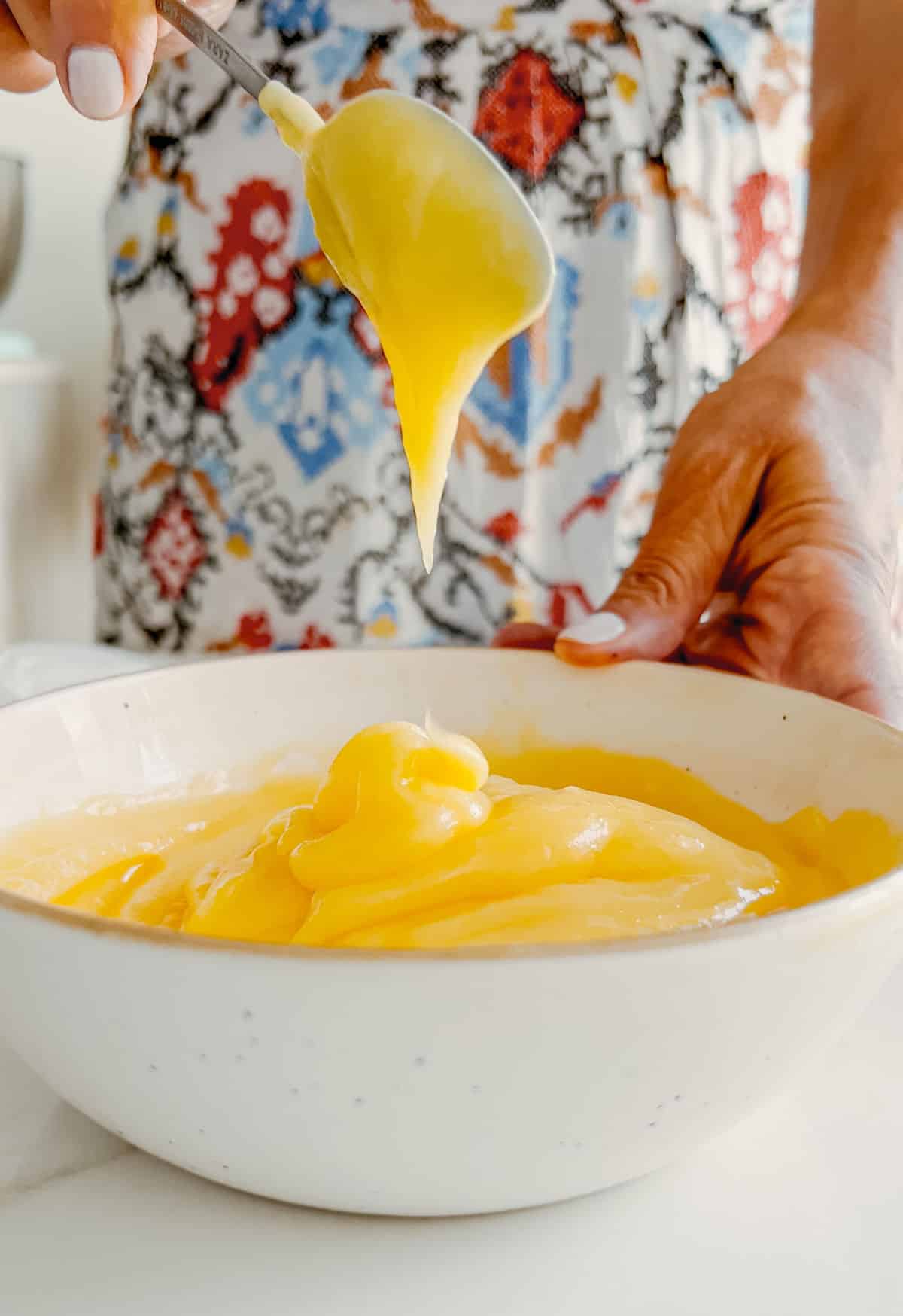 White bowl with lemon curd being spooned by person with colorful apron.