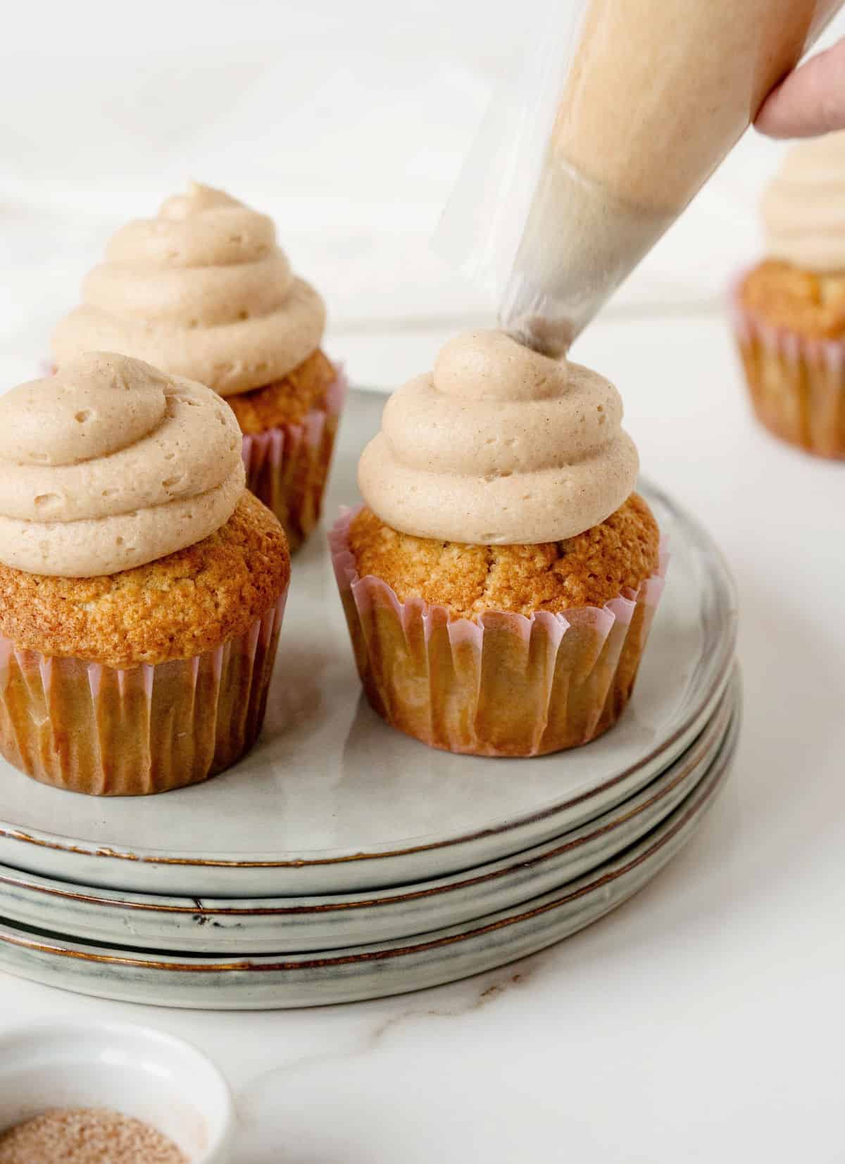 Using a piping bag to frost cinnamon cupcakes in paper liners on a stack of grey plates. White surface and background.