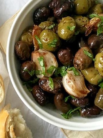 Partial close up view of roasted olives and garlic in a white bowl with bread pieces beside it on a white cloth.