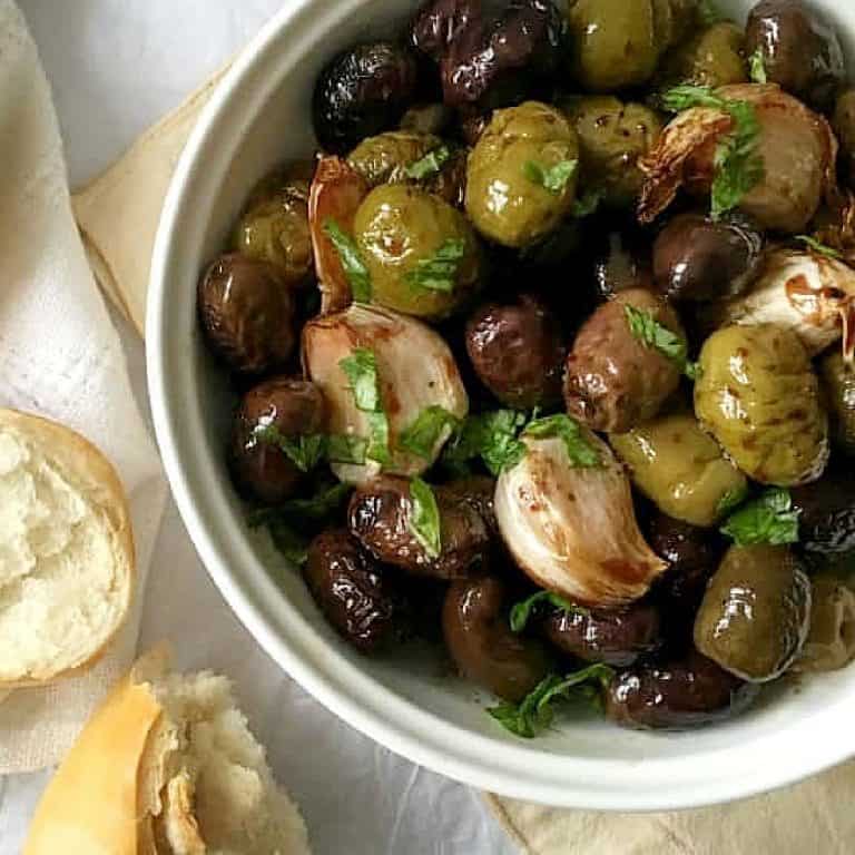 Partial close up view of roasted olives and garlic in a white bowl with bread pieces beside it on a white cloth.