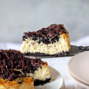 Single slice of Oreo cheesecake being lifted on a metal cake server. Whole cheesecake below. Grey and white background.