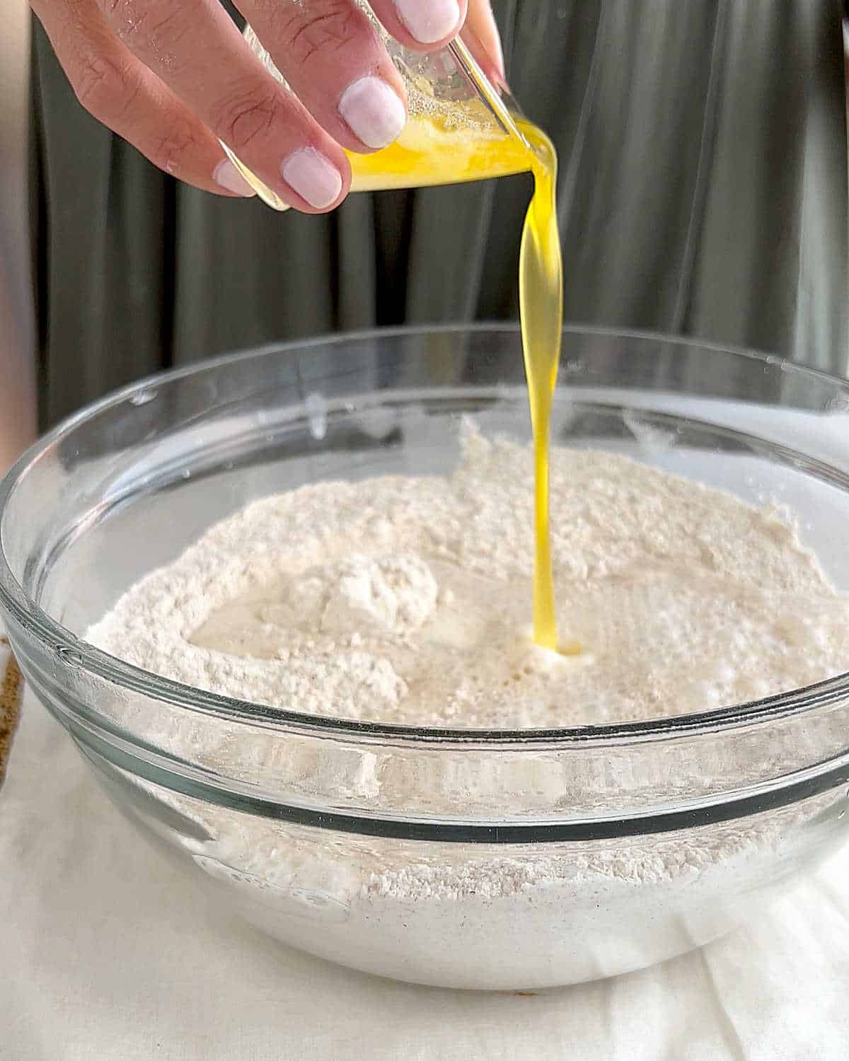 Hands adding melted butter to whole wheat mixture in a glass bowl. White surface, green background.
