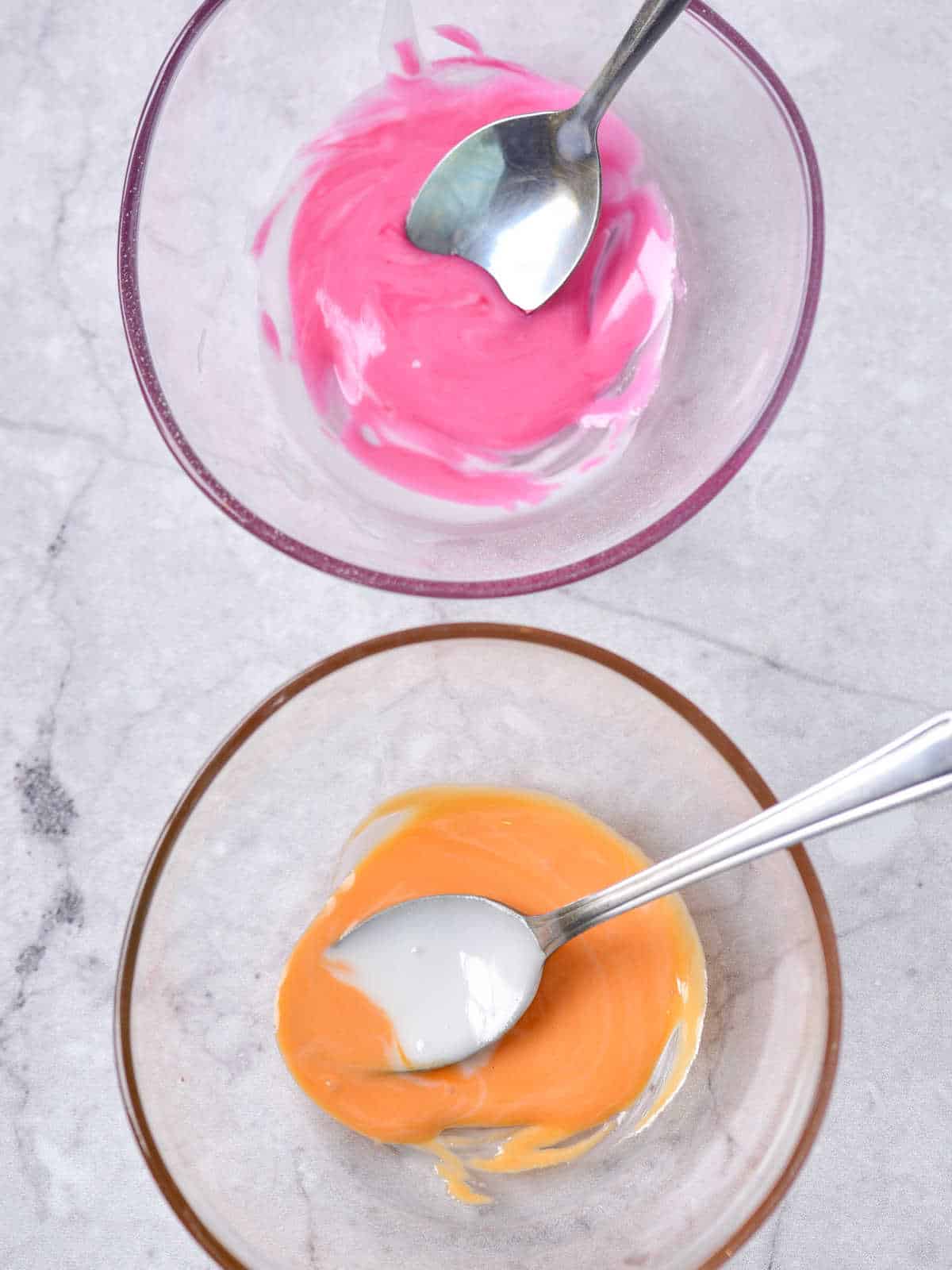 Two glass bowls with spoons on a grey surface with orange and bright pink icing.