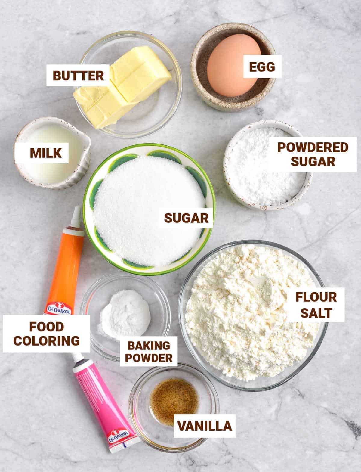 Grey surface with bowls containing ingredients for sugar cookies including egg, butter, food coloring, vanilla, sugars, flour mixture.