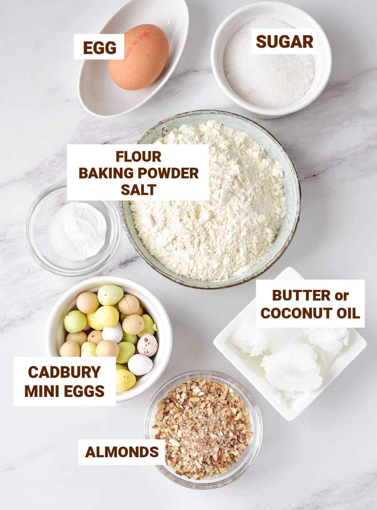 White marble with bowls containing ingredients for Cadbury egg cookies including coconut oil, almonds, flour mixture, egg, sugar.