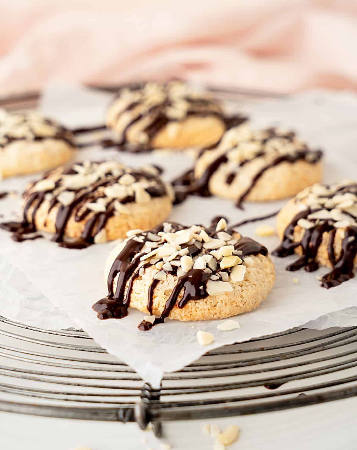 Several chocolate drizzled almond cookies on parchment paper set on a wire rack. Pink background.