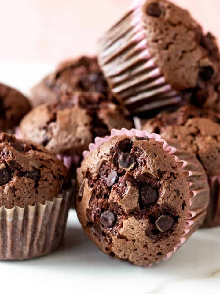 A pile of chocolate muffins in paper liners on a white surface with a pink background.