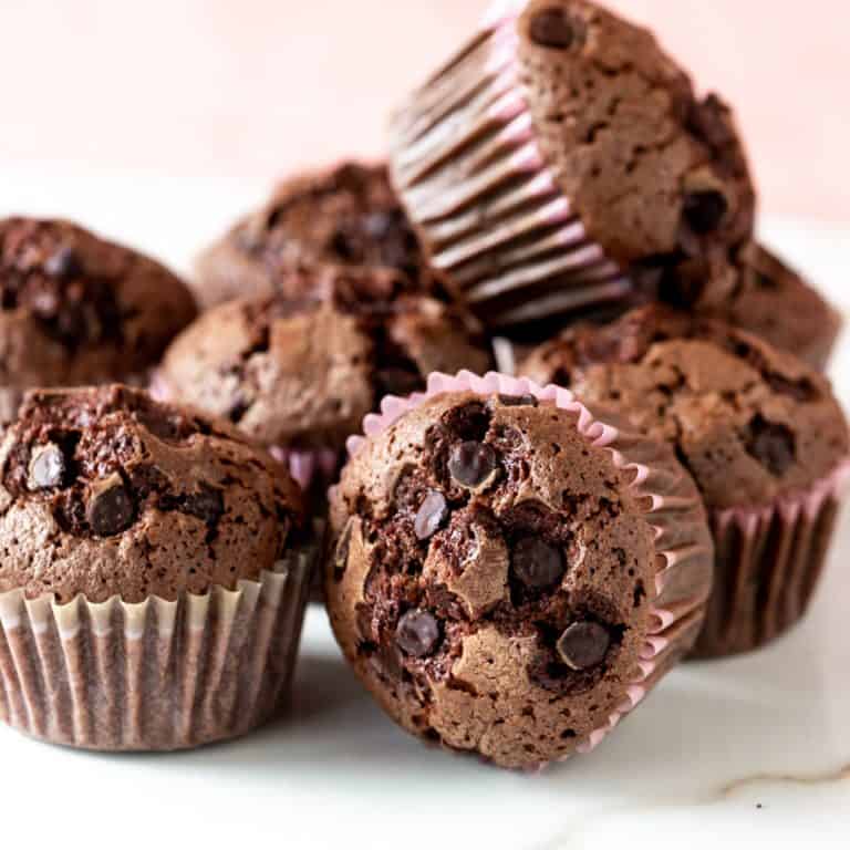 A pile of chocolate muffins in paper liners on a white surface with a pink background.