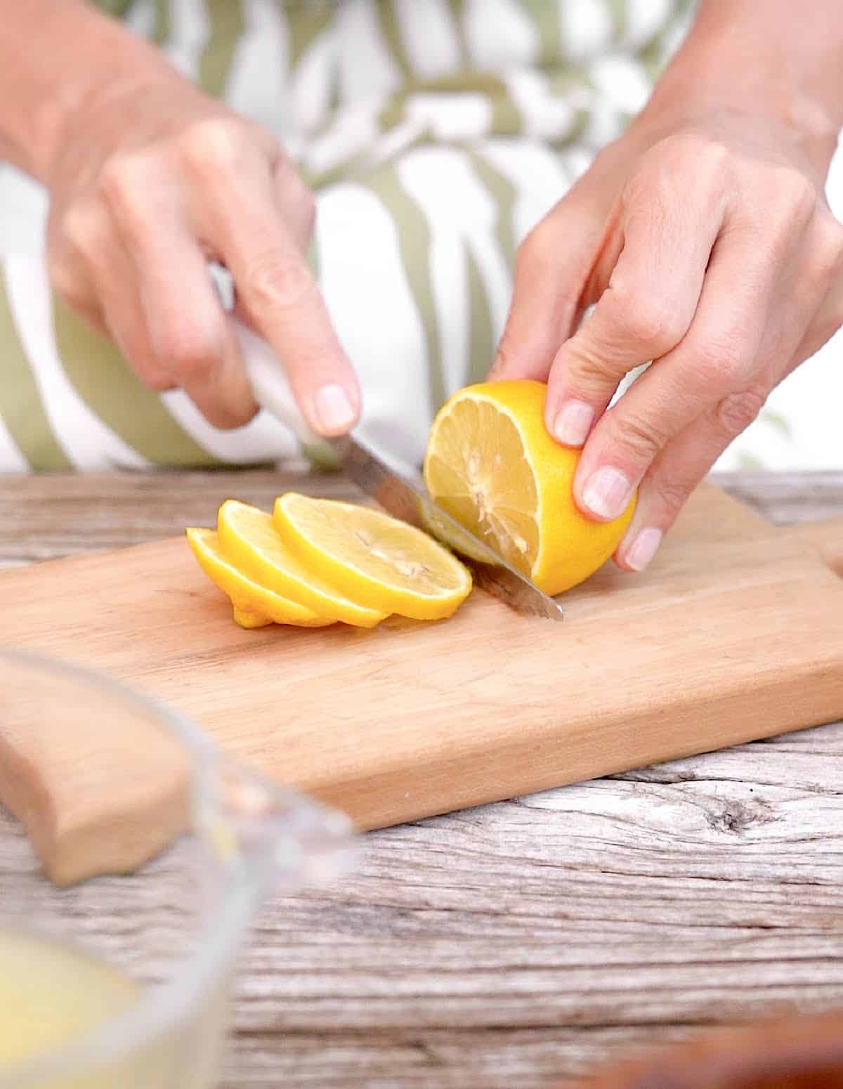 Cutting lemon slices on a light colored wooden board. White and green dress in the background.