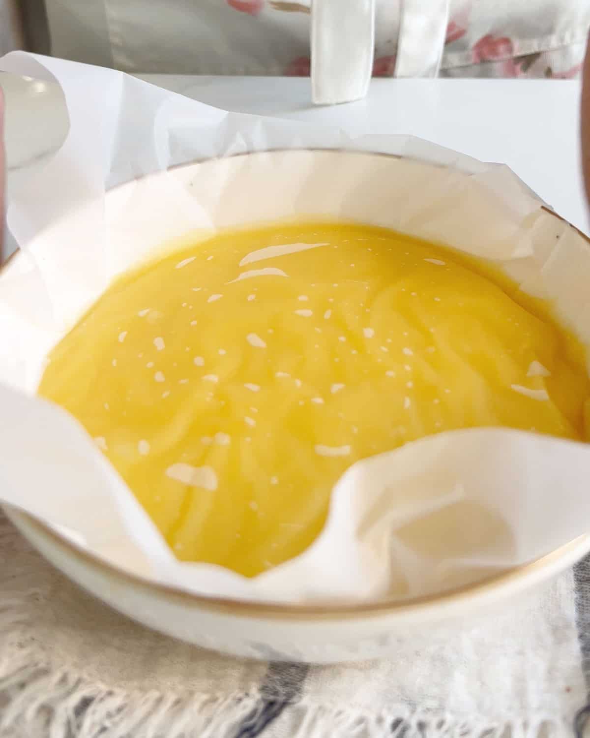 Piece of plastic covering lemon curd in a white bowl on a white surface with a striped cloth. 