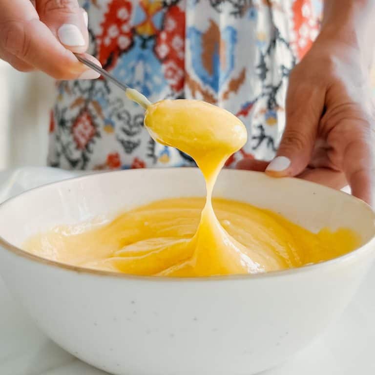 Lifting a spoon full of lemon curd from a white bowl. Colorful cloth as background.