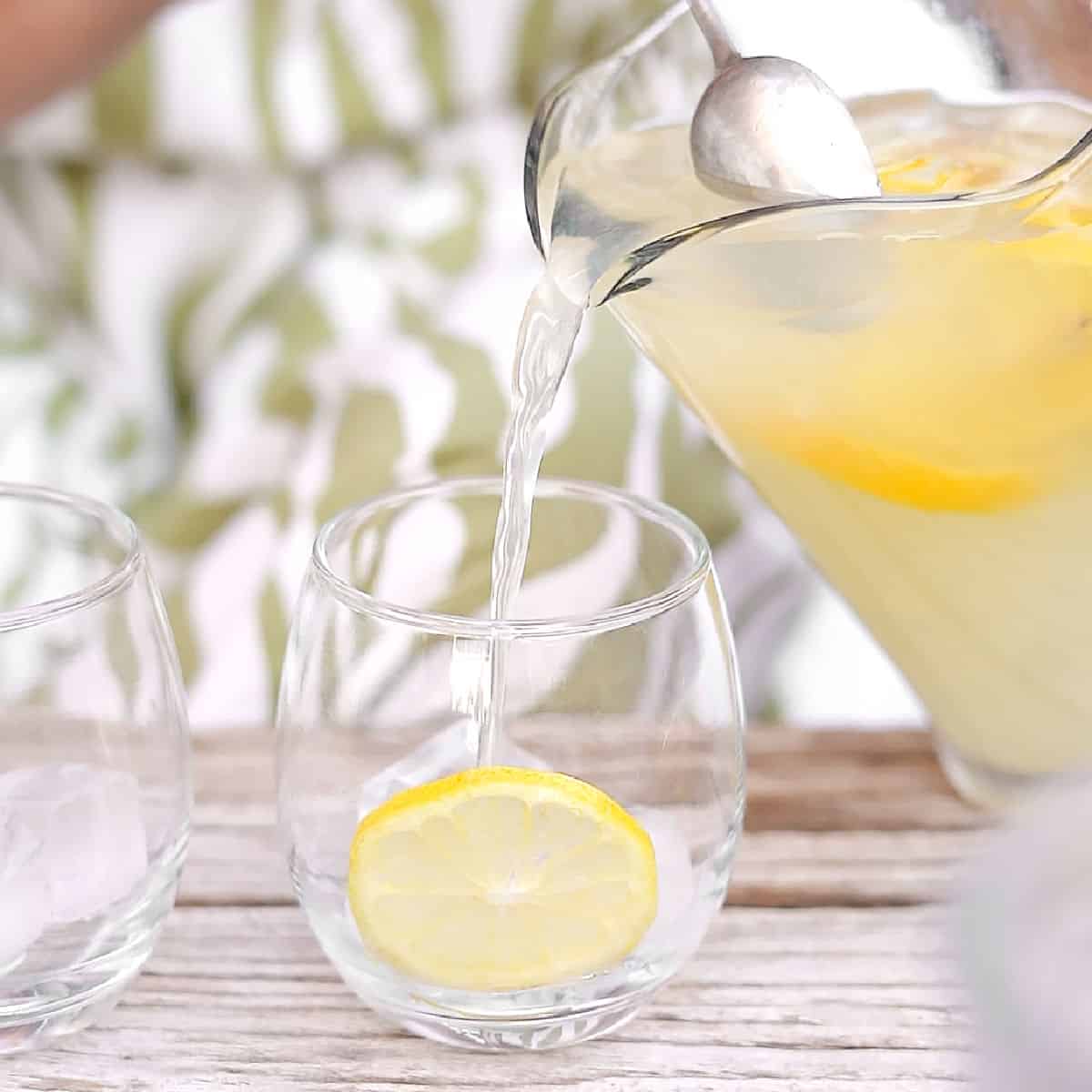 Serving lemonade from a jar into a glass with lemon slices. Wooden table. Green and white background.