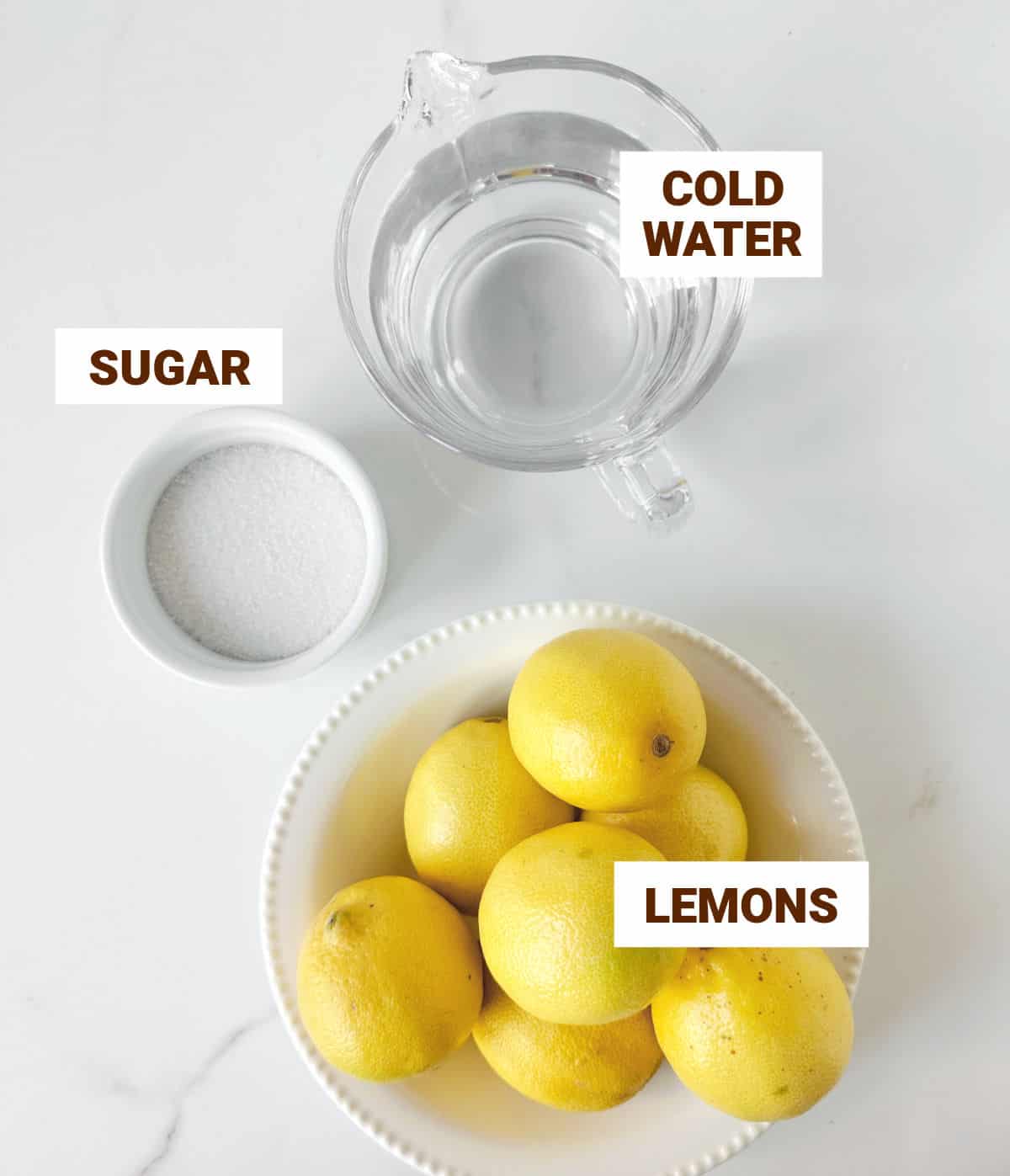 White marble surface with ingredients for lemonade including pitcher of water, whole lemons, and sugar in a white bowl.