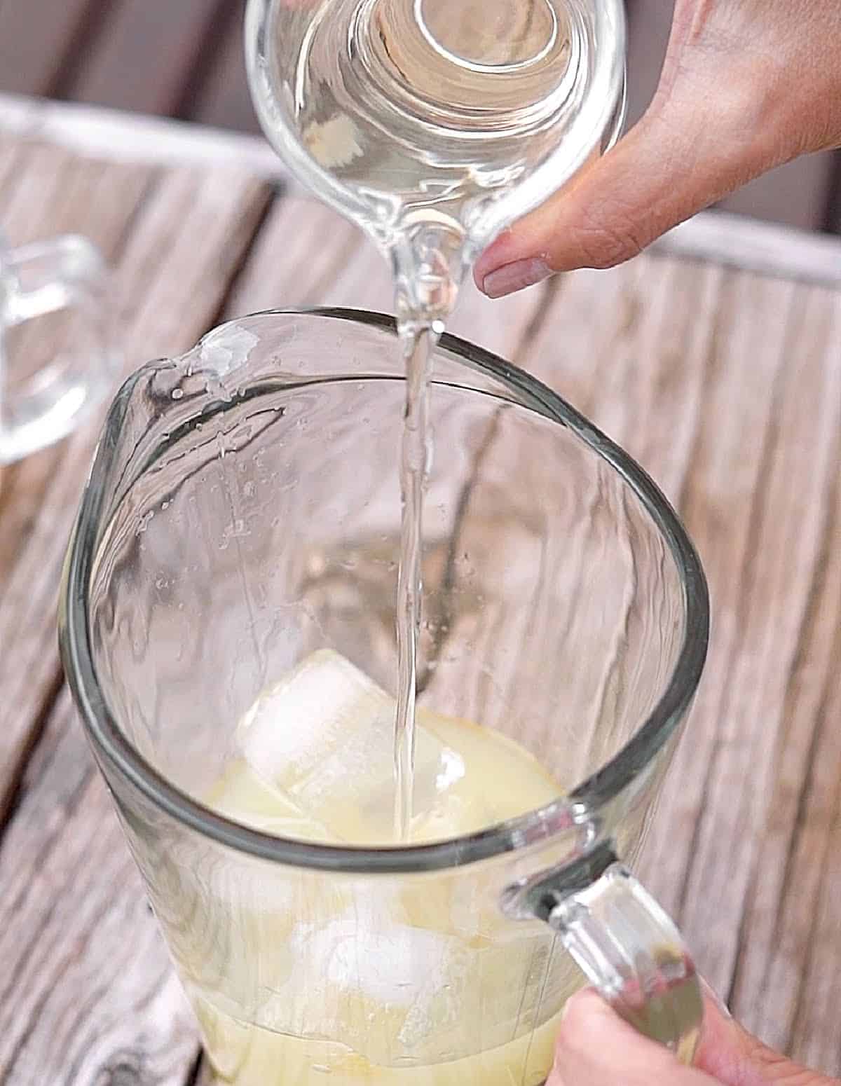 Syrup being added to a pitcher with lemon juice and ice cubes. Grey wooden surface.