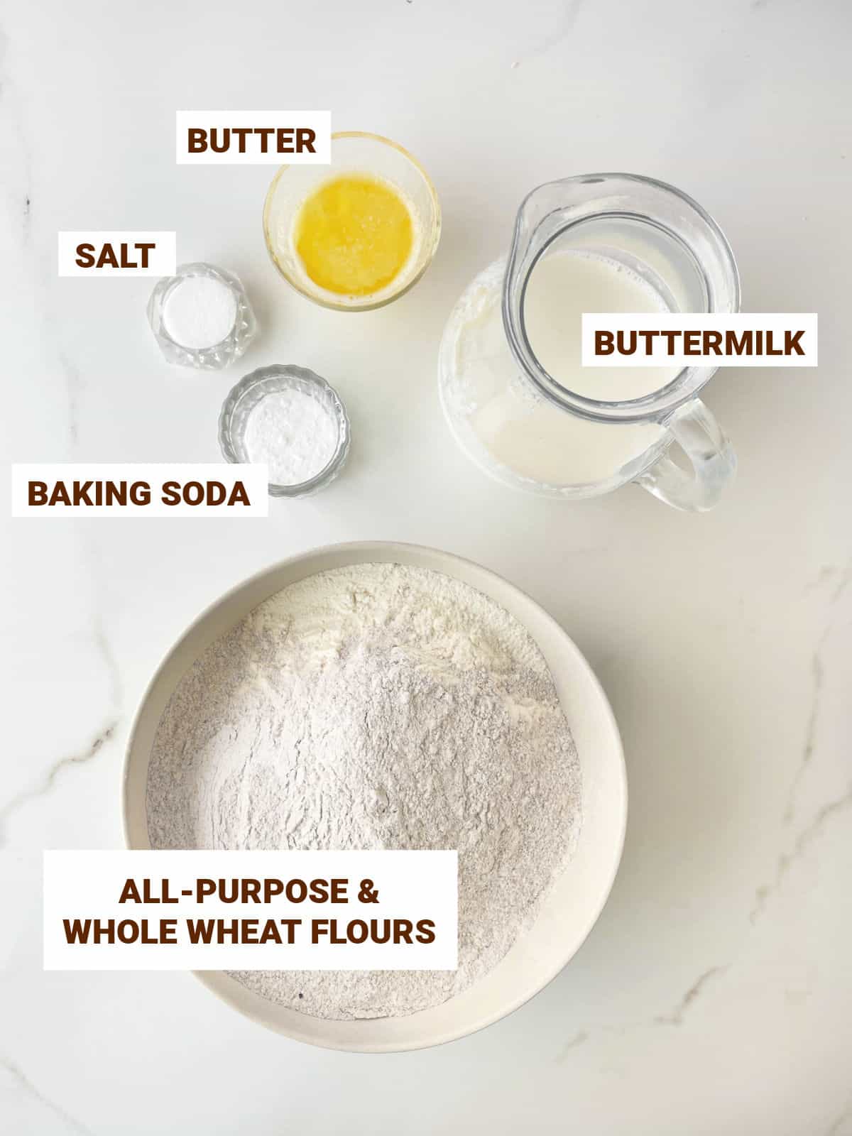 Ingredients for whole wheat soda bread in bowls on a white marble surface including butter, flours, salt, and buttermilk.