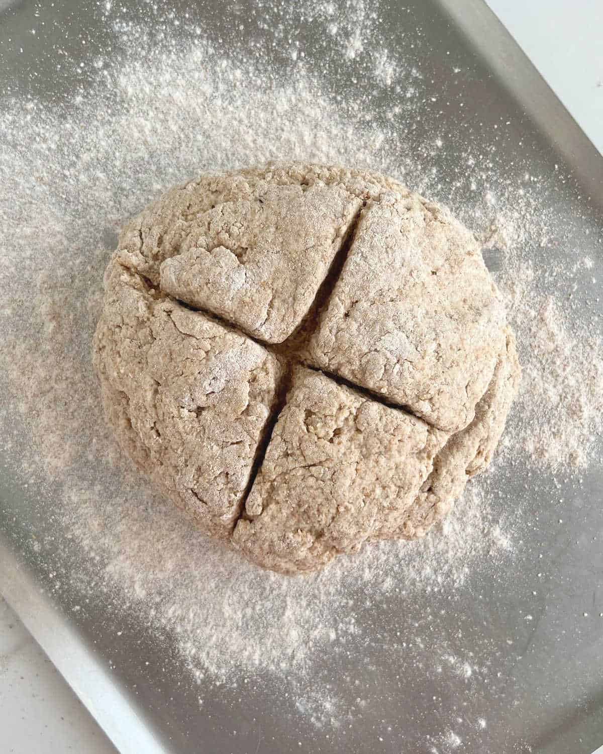 Round loaf of whole wheat bread dough with a cross on top on a floured metal baking sheet.