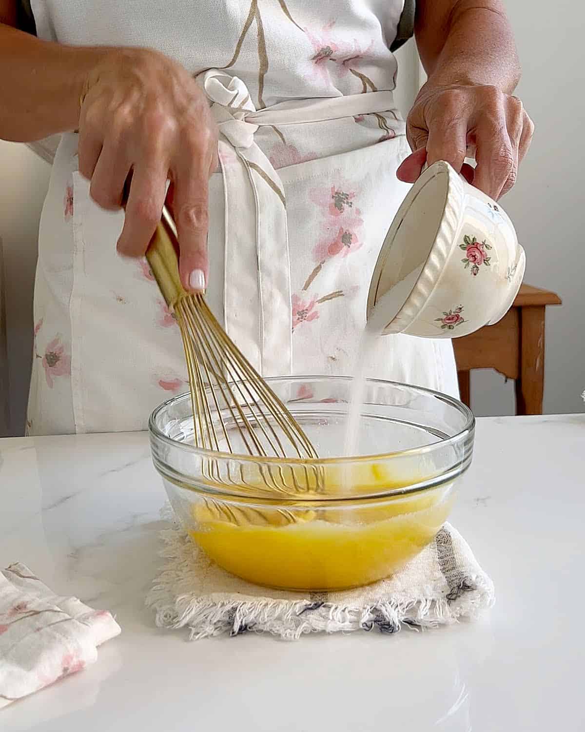 Adding sugar from an old-fashioned cup into egg mixture in a glass bowl. Gold whisk.
