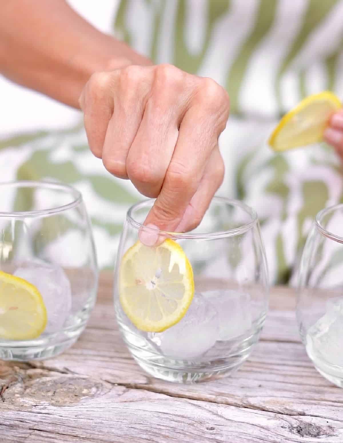 Hand placing lemon slices inside glasses on a greyish wood table. Green and white dress as background.