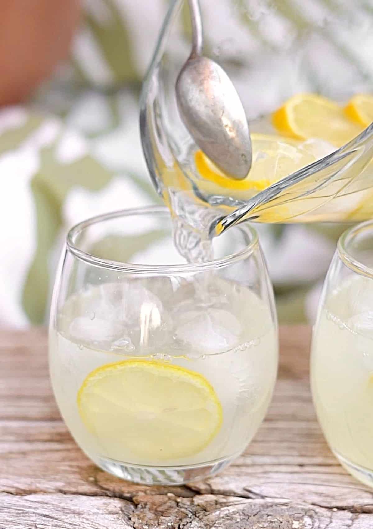 Homemade lemonade being served from a jar into a glass with lemon slice on a wooden surface. Green and white background.
