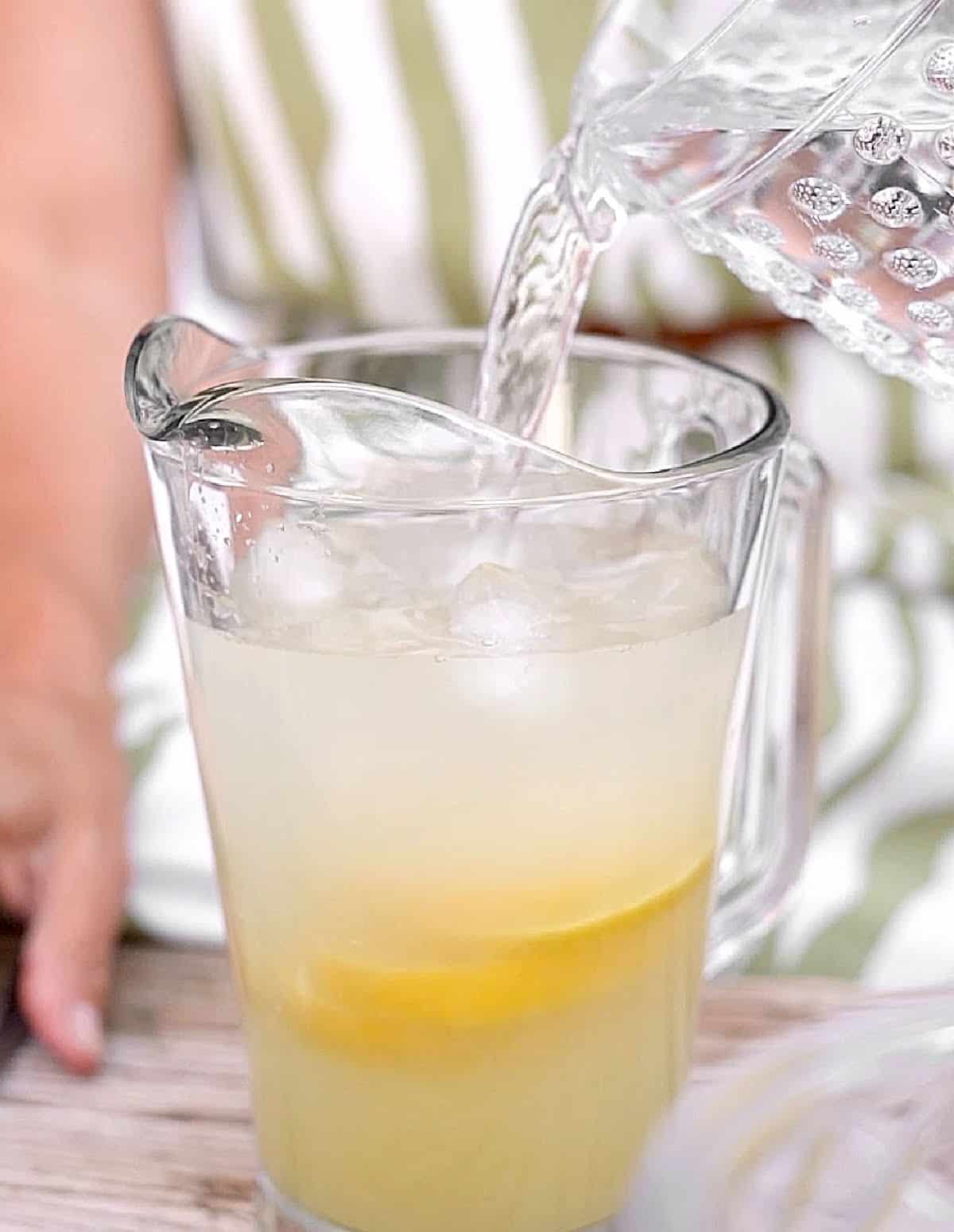 Water being added to a large pitcher with lemonade and lemon slices.