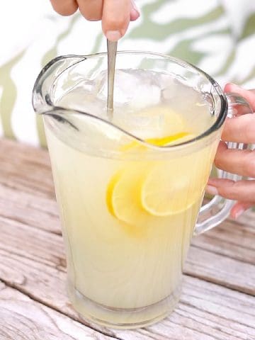 Jar of lemonade with lemon slices being stirred on a light wooden table.